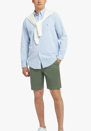 Blauw-wit gestreepte button-up shirt, groene shorts, witte trui om de schouders gebonden, en witte sneakers.