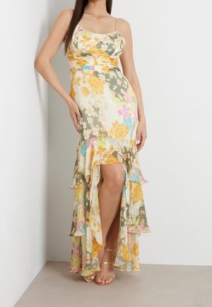 Femme portant une robe fleurie sans manches avec un ourlet volanté haut-bas et des talons dorés à brides, debout contre un mur blanc.
