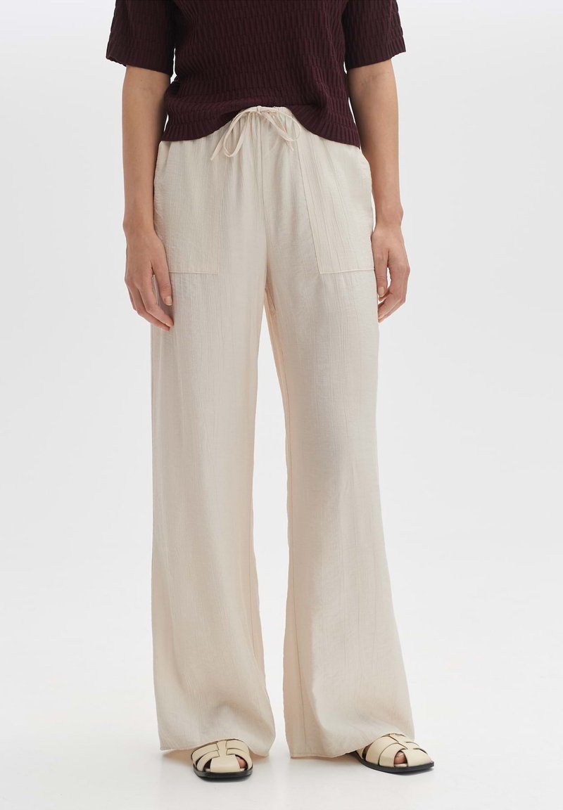 Lichtbeige wijde broek met twee voorkezen en een trekkoord in de taille, gemaakt van een zachte textuur. Gecombineerd met zwarte spiked sandalen.