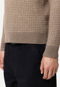 Beige texturerad stickad tröja med ribbad nederkant och muddar. Bärs med mörka byxor, vilket framhäver subtila färgkontraster och materialdetaljer.