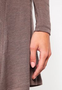 Hnědý pletený svetr s dlouhými rukávy a texturovaným vzorem. Ruka umístěná blízko lemu, ukazující měkký materiál a barvu.