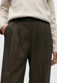 Braune maßgeschneiderte Hosen mit Falten, die eine glatte Textur und einen klassischen Schnitt aufweisen. Getragen mit einem hellbeigen Pullover.