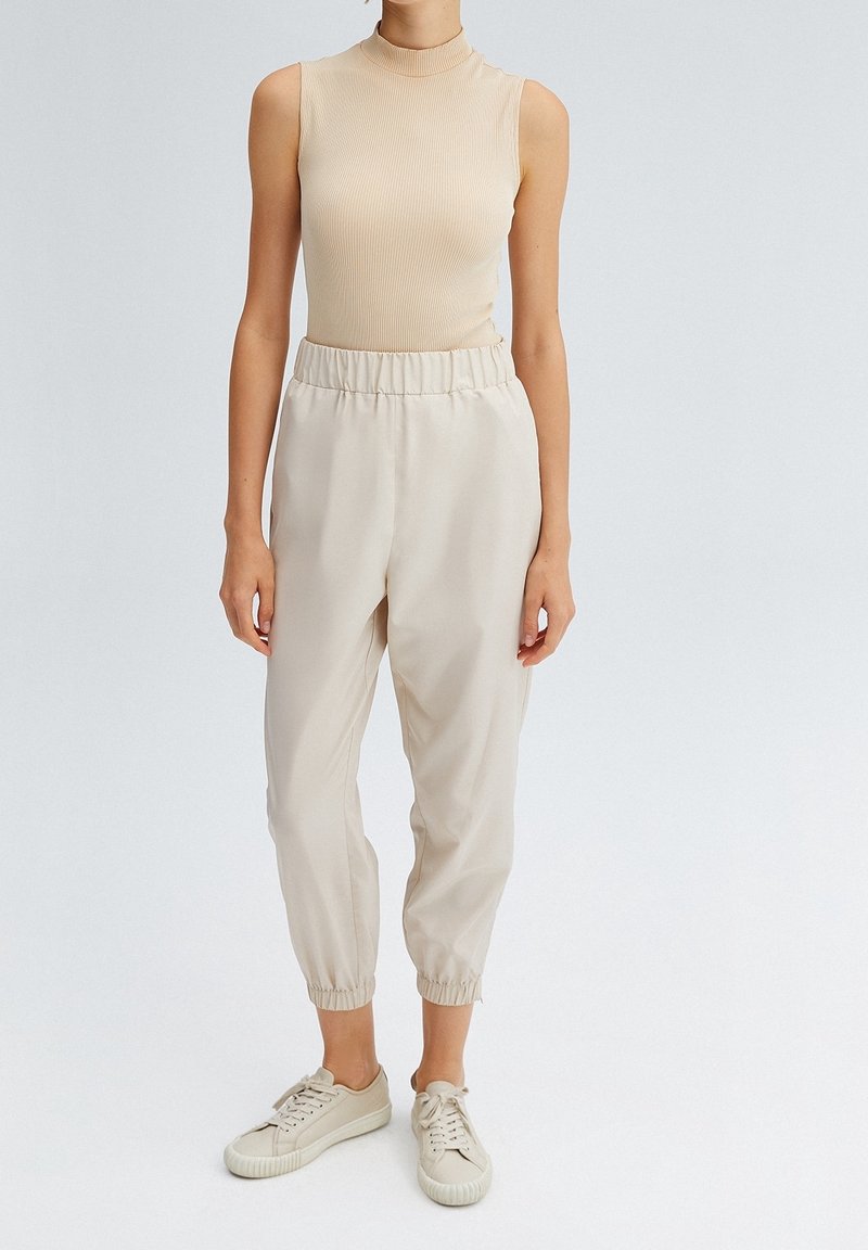 Beige geribbelde mouwloze top gecombineerd met lichte, crèmekleurige, taperende broek met een elastische tailleband en opgerolde enkels, afgemaakt met beige sneakers.