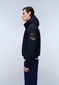 Młody mężczyzna w granatowej kurtce z kapturem, z kieszenią na zamek i naszywkami z logo na rękawie, stojący bokiem na jednolitym tle.