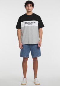 Trumpas rankovėmis marškinėliai su juodais, baltais ir pilkais spalvų blokais, ant kurių juodu raštu užrašyta "GUESS JEANS SINCE 1981". Suderinta su džinsiniais šortais.