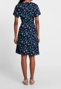 Tummansininen mekko, jossa on lyhyet hihat, vihreä- ja sinikukkakuvio, vyötä ympäröivä istuvuus, virtaava hame ja teksturoitu kangas.