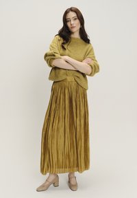 Olivgrüner gestrickter Pullover kombiniert mit einem plissierten, knielangen goldenen Rock. Tanfarbene Schuhe mit Blockabsatz runden das Outfit ab.