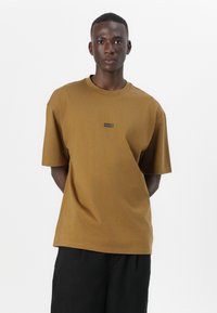 Brun bomulls t-skjorte med korte ermer, med en svart rektangulær "HUGO" logo på brystet, avslappet passform og rund hals.