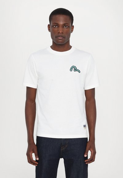 T-shirt en coton blanc à manches courtes, avec un design abstrait vert sur la poitrine gauche et une petite étiquette logo en bas.