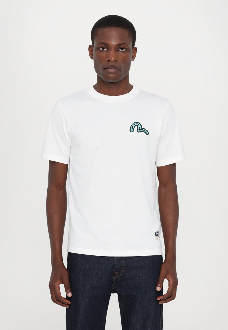 Camiseta de algodón blanca de manga corta, con un diseño abstracto verde en el lado izquierdo del pecho y una pequeña etiqueta de logo en el dobladillo.