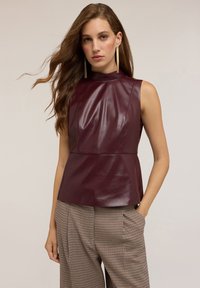 Donna con lunghi capelli castani che indossa lunghe orecchie d'oro, un top in pelle bordeaux senza maniche e pantaloni a scacchi marroni, in piedi con una mano in tasca.