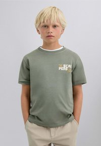 Camiseta de algodón verde con mangas cortas, que presenta un pequeño estampado gráfico en el lado izquierdo del pecho. Corte casual, diseño de cuello redondo. Textura suave.
