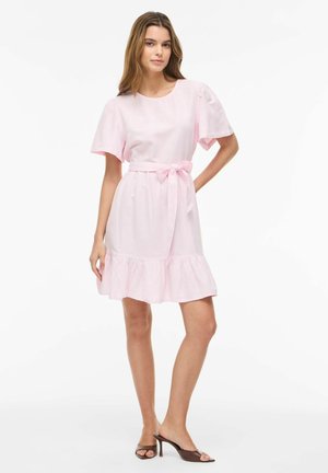 Kobieta stojąca, ubrana w jasnoróżową sukienkę z krótkim rękawem, związaną paskiem i falbaną na dole, w połączeniu z brązowymi sandałami na obcasie z otwartymi palcami.