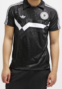 Černý sportovní dres s texturovaným povrchem, zdobený bílým chevron vzorem, kulatým logem a třemi bílými pruhy na rukávech.