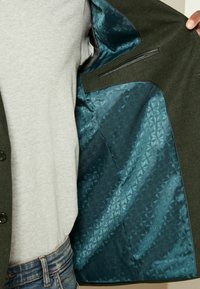 Olivgrüne Jacke mit einer strukturierten Außenseite, die ein glattes türkisfarbenes Satinfutter mit einem geometrischen Muster und einem versteckten Taschen-Detail aufweist.
