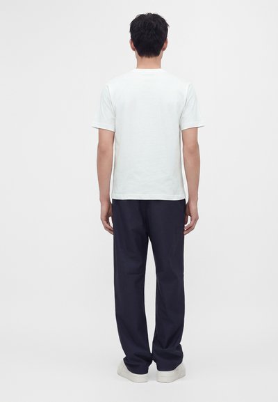 Άνδρας με σκούρα μαλλιά, φοράει απλό λευκό t-shirt με κοντά μανίκια, σκουρόχρωμο παντελόνι και λευκά παπούτσια, στέκεται γυρισμένος με την πλάτη σε λευκό φόντο.