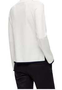 Person som bär en vit långärmad topp med svarta detaljer och svarta byxor, visad bakifrån med händerna i fickorna.