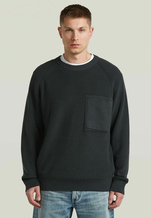 Giovane uomo con capelli corti che indossa un maglione scuro con taschino sul petto e jeans azzurri, in piedi davanti a uno sfondo chiaro e semplice.