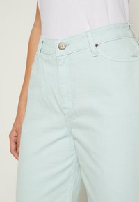 Jeans de ganga verde menta claros com um design clássico de cinco bolsos, fecho com botão metálico e costuras detalhadas na parte da frente.