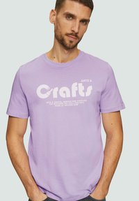 Paarse katoen t-shirt met een witte geprinte opdruk die "Arts & Crafts" zegt, vergezeld van grafische accenten. Korte mouwen en een ronde hals.