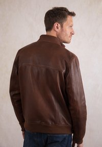 Man turning sideways wearing a brown leather jacket and blue jeans against a plain light background.