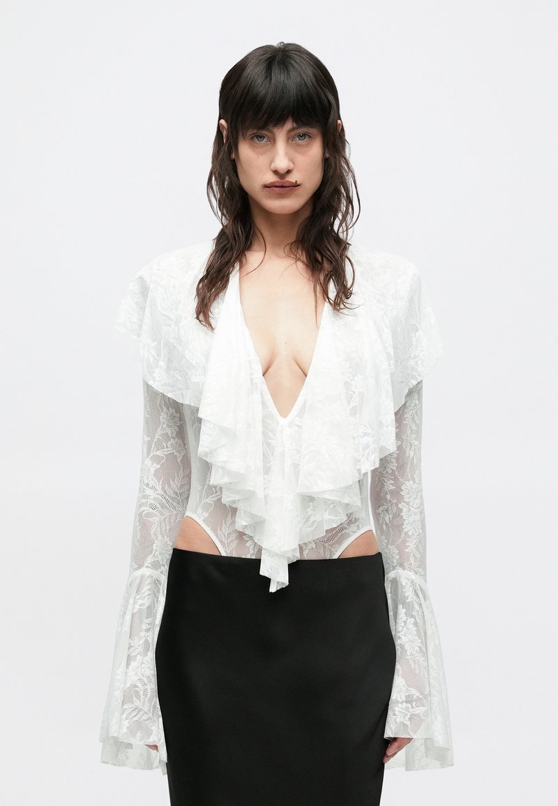 Γυναίκα με μακριά σκούρα μαλλιά φοράει λευκό δαν�τελένιο πουκάμισο με βαθύ V και βολάν, συνδυασμένο με μαύρη ψηλόμεση φούστα, στέκεται μπροστά σε απλό φόντο.