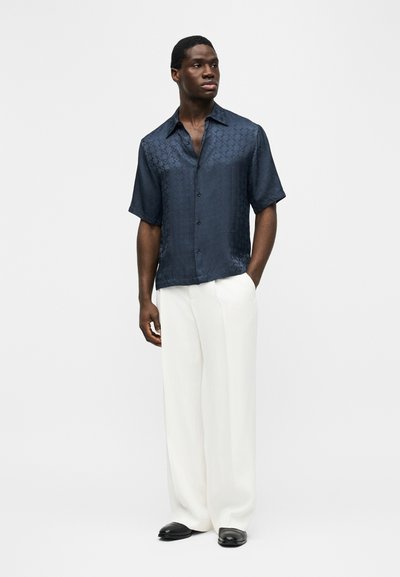 Άνδρας που φοράει σκούρο μπλε πουκάμισο με κοντά μανίκια και λεπτό μοτίβο, καθώς και πλατύ λευκό παντελόνι, στέκεται με το ένα χέρι στην τσέπη.