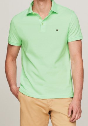 Mann trägt hellgrünes kurzärmeliges Poloshirt mit kleinem Logo auf der Brust und beige Hose, Hand in der Tasche, neutraler Hintergrund.