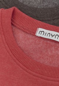 Dwa dekolty t-shirtów nachodzące na siebie: jeden wykonany z teksturowanej czerwonej bawełny z ribbowanym wykończeniem i metką, drugi z gładkiej ciemnoszarej bawełny.