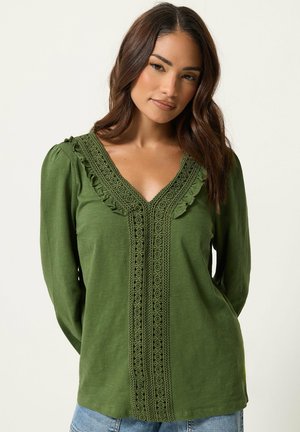 Mujer con cabello largo castaño que lleva una blusa verde con encaje y volantes a lo largo del escote en V y el centro delantero, combinada con vaqueros azul claro.