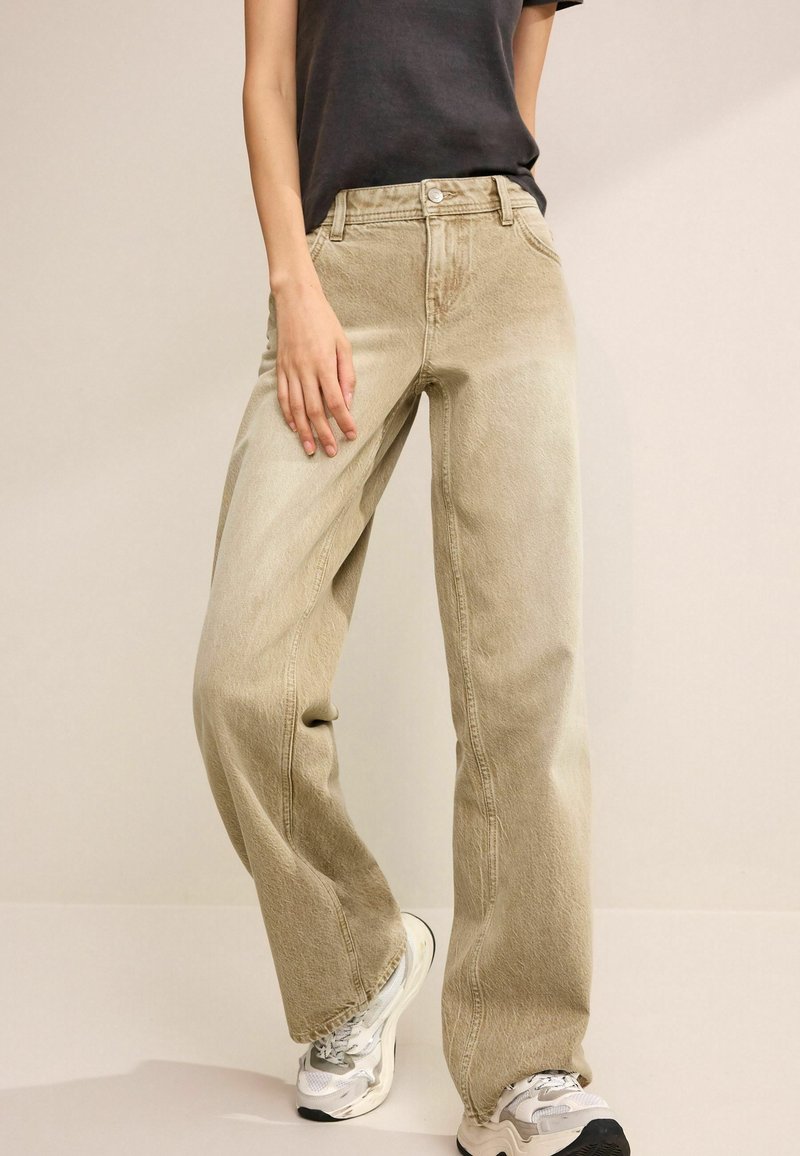 Personne portant un jean beige ample, un t-shirt noir et des baskets blanches, se tenant contre un fond neutre uni.