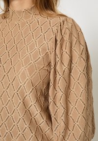 Beige bluse med struktur og høj hals, prydet med et diamantmønster og subtil åben vævning. Lange ærmer med detaljerede samlinger.