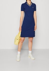 Navyblaues Polokleid mit V-Ausschnitt und gestreiftem Kragen, aus weichem Stoff. Kombiniert mit einer gelben Daunen-Tasche und weißen Sneakers.