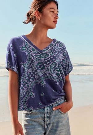 Ung kvinde på stranden iført en løs lilla top med paisley-mønster og lyseblå jeans med den ene hånd i lommen, kigger til siden.