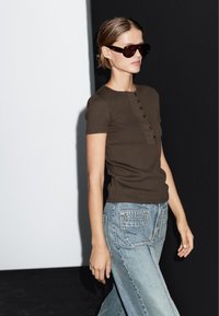 Hnědý žebrovaný top s krátkým rukávem, kulatým výstřihem a detaily knoflíků, v kombinaci se světle modrými širokými džíny s prošitím zadních kapes.