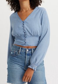 Top lungo azzurro chiaro a maniche lunghe con scollatura a V, chiusura con bottoni e vita arricciata. Abbinato a pantaloni shorts in denim usurato a vita alta.