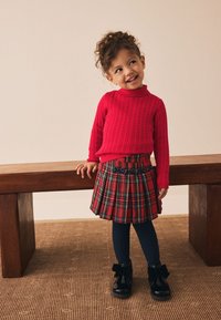 Piros kötött pulóver, kockás pliszírozott szoknya tengerészkék részletekkel, tengerészkék kötött harisnya és fekete patent bőr bokacsizma masnikkal.