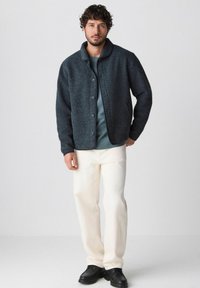 Giacca di lana grigia scura con bottoni frontali, colletto grande, indossata sopra una camicia teal, abbinata a pantaloni color crema e scarpe nere.