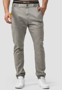 Graue Jogginghose mit schmalem Schnitt, ausgestattet mit einer subtilen Textur, elastischen Bündchen und einem schwarzen Bund mit "INDICODE"-Logo. Gelbes Etikett sichtbar.