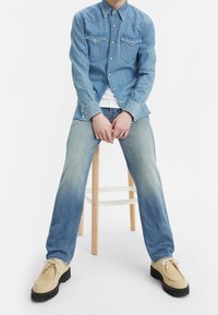 Persona sentada en un taburete de madera, vistiendo una camisa de mezclilla azul claro, unos jeans azules desteñidos, zapatos beiges con cordones y una camiseta blanca debajo.