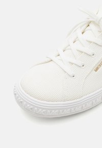 Baskets blanches avec tige en mesh, lacets blancs plats et détail de logo doré. Semelle blanche texturée arborant un motif répétitif de "MK."