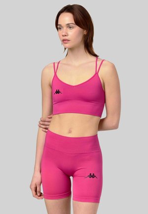 Femme portant un brassière de sport rose et un short assorti avec des logos noirs, debout devant un fond gris clair uni.