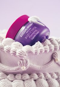 Fioletowy szklany słoik z etykietą "EQUALBERRY DEEP CERA BAKUCHIOL Krem w kapsułkach nawadniający" spoczywa na teksturowanej, pastelowej powierzchni z lukrem ozdobionym spiralkami.