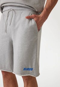 Pantaloni corti grigi in cotone con vita elasticizzata, tasche laterali e un logo blu "GUESS" sull'orlo. Finitura testurizzata con bordi sfilacciati.