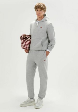 Joven con sudadera con capucha gris claro y pantalones deportivos, sosteniendo una bolsa a rayas roja y blanca, de pie frente a un fondo plano y claro.