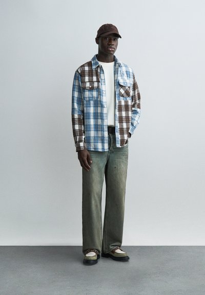 Άνδρας με καφέ καπέλο, μπλε και καφέ καρό πουκάμισο πάνω από λευκό μπλουζάκι, ξεθωριασμένο πράσινο τζιν και δίχρωμα παπούτσια στέκεται μπροστά σε απλό φόντο.