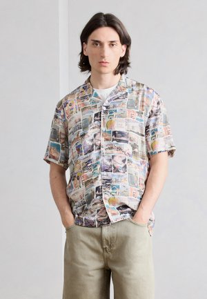 Νέος άνδρας με μαλλιά μήκους έως τους ώμους φορά πολύχρωμο πουκάμισο με κοντά μανίκια και εκτυπώσεις ταξιδιωτικών καρτ ποστάλ και ανοιχτό μπεζ παντελόνι.