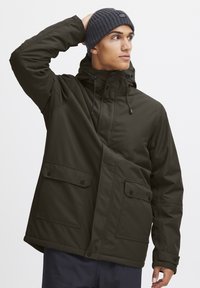 Giacca bicolore verde scuro con zip e chiusura a scatto, cappuccio e tasche sul petto. Presenta una texture liscia e cordini regolabili.