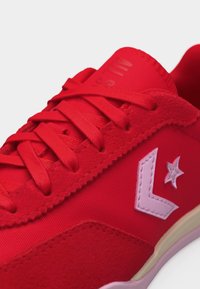 Κόκκινο αθλητικό παπούτσι με κορδόνια στο ίδιο χρώμα, λευκή σειρά σε σχήμα βέλους και λογότυπο αστέρι στο πλάι, καθώς και σουέντ λεπτομέρειες κοντά στην περιοχή των δαχτύλων.