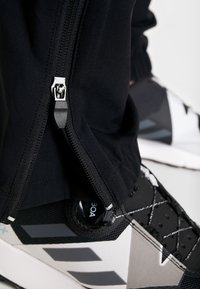 Pantaloni sportivi neri con cerniera laterale e finitura texturizzata, abbinati a scarpe sportive nere e grigie con parte superiore in rete e sistema di lacci.
